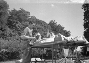 18th Field Ambulance medical assistant offers a drink to patient Bourgtheroulde, France, 26 August 1944. Source: TBC